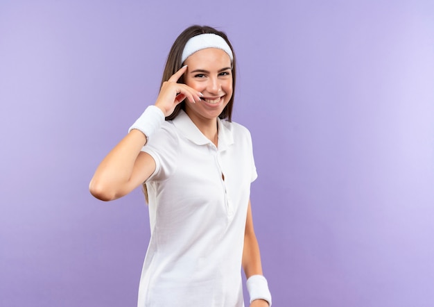 Garota muito esportiva sorridente usando bandana e pulseira colocando o dedo na têmpora isolada no espaço roxo