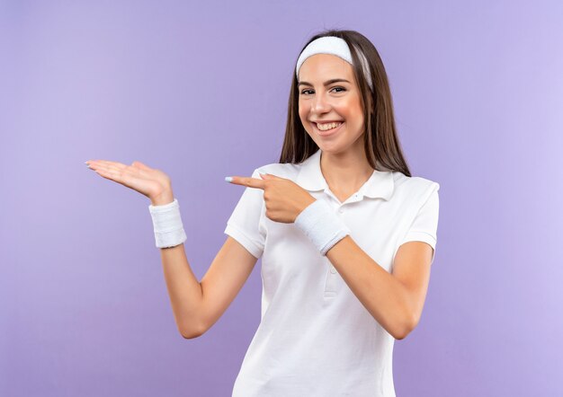 Garota muito esportiva sorridente usando bandana e pulseira, mostrando a mão vazia e apontando para ela isolada no espaço roxo