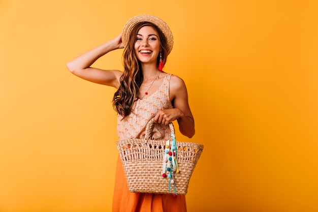 Garota muito glamourosa tocando seu chapéu de palha. Retrato de estúdio de jovem alegre com bolsa de verão.