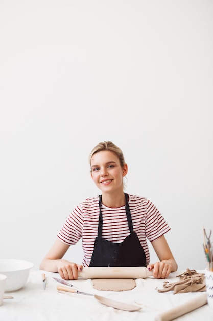 Garota muito sorridente de avental preto e camiseta listrada, sentado à mesa com rolo e trabalhando com argila alegremente olhando na câmera no estúdio de cerâmica
