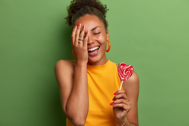 Foto grátis garota negra alegre faz cara de palma, sorri amplamente, fecha os olhos, posa com pirulito de coração no palito, se diverte em casa, segura doces deliciosos, usa camiseta amarela, fica de pé contra a parede verde vívida