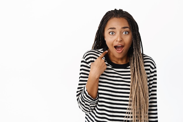 Garota negra surpresa apontando para si mesma com descrença e felicidade ganhando algo sendo escolhido em pé sobre fundo branco