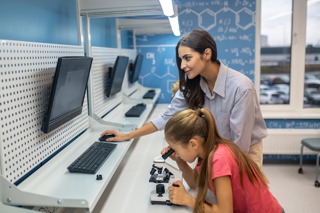 Garota olhando através do microscópio e professor em pé nas proximidades