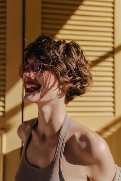 Garota otimista com penteado encaracolado rindo no pano de fundo amarelo Mulher legal em óculos de sol rosa posa no fundo brilhante