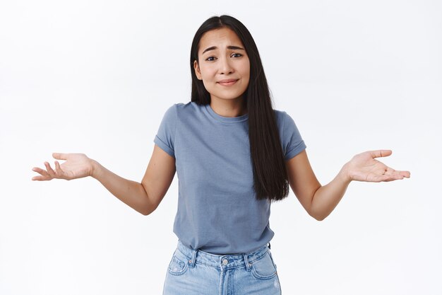 Garota pedindo desculpas, não consigo decidir ou pensar em uma solução, encolhendo os ombros e estendendo as mãos para os lados sem esperança, sorrindo com uma expressão inocente de desculpas, sendo insegura ou duvidosa, não posso dizer, parede branca