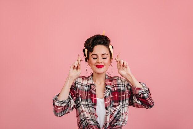 Garota Pin-up posando com os olhos fechados e dedos cruzados. Senhora caucasiana alegre em pé de camisa quadriculada no fundo rosa.