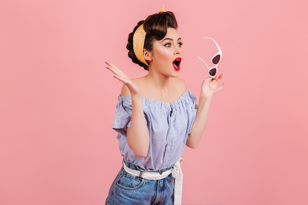 Foto grátis garota pinup chocada segurando óculos de sol. foto de estúdio de emocional mulher com roupa vintage isolada no fundo rosa.