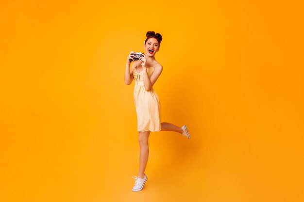 Garota pinup emocional com câmera dançando em fundo amarelo Foto de estúdio de fotógrafa feminina de vestido