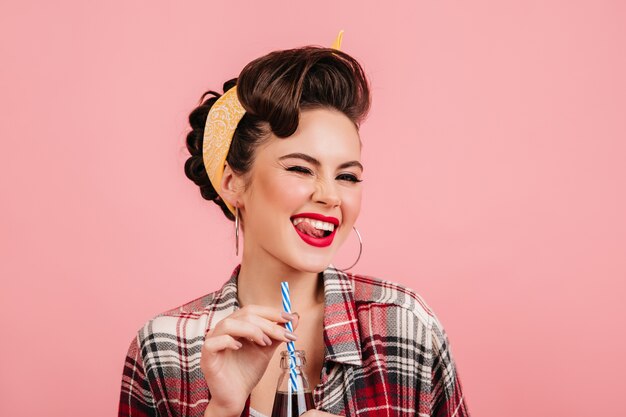 Garota pinup sonhadora com maquiagem brilhante, rindo no fundo rosa. Foto de estúdio de senhora despreocupada bebendo bebida.