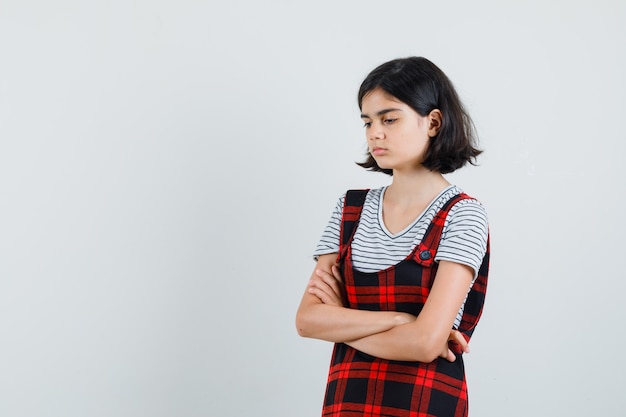 Garota pré-adolescente em pé com os braços cruzados em t-shirt, macacão e olhando ofendida, vista frontal. espaço para texto