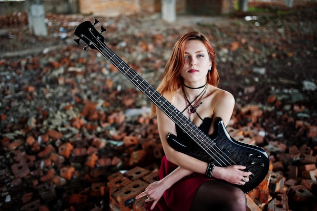 Foto grátis garota punk de cabelo vermelho usa saia preta e vermelha com guitarra baixo em lugar abandonado retrato de músico gótico