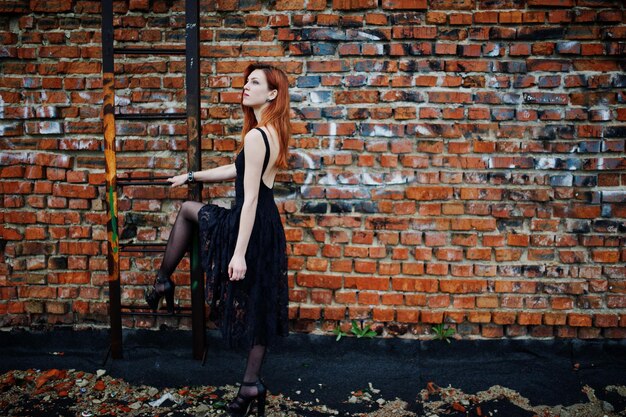 Garota punk de cabelos vermelhos usa vestido preto no telhado contra a parede de tijolos com escada de ferro