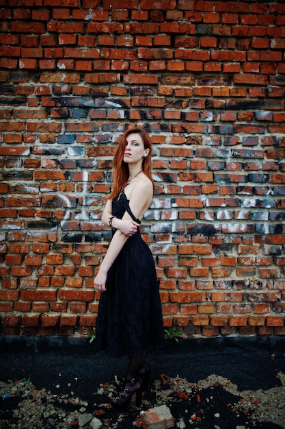 Garota punk de cabelos vermelhos usa vestido preto no telhado contra a parede de tijolos com escada de ferro