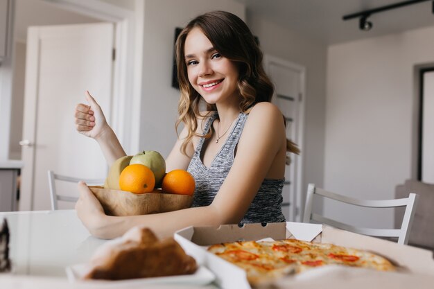 Garota relaxada, abraçando o prato com frutas. Mulher encaracolada glamorosa posando com laranjas e rindo.