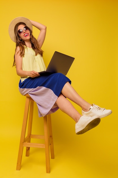 Garota relaxada usando um vestido de verão brilhante, trabalhando com um laptop, sentada na cadeira sobre uma parede isolada