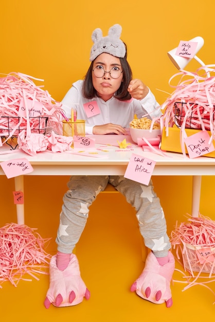 Foto grátis garota repreende você aponta para a câmera com expressão irritada vestida de pijama senta na mesa do escritório com pilhas de papel cortado faz adesivos de memorando trabalhos de casa isolados na parede amarela