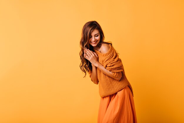 Garota romântica com penteado ondulado, posando em laranja com um sorriso. Retrato interior da incrível senhora branca de suéter da moda.