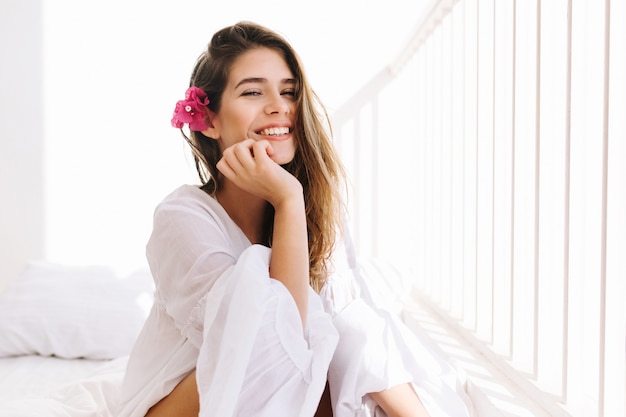 Garota romântica com um sorriso malicioso na blusa vintage, sentada na cama e tocando seu queixo com a mão. Retrato de uma jovem fofa sonhadora com flor no penteado, descansando no quarto pela manhã