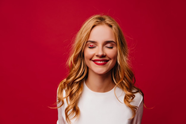 Garota romântica de cabelos compridos posando com um sorriso tímido. Foto interna de atraente mulher europeia isolada na parede vermelha.