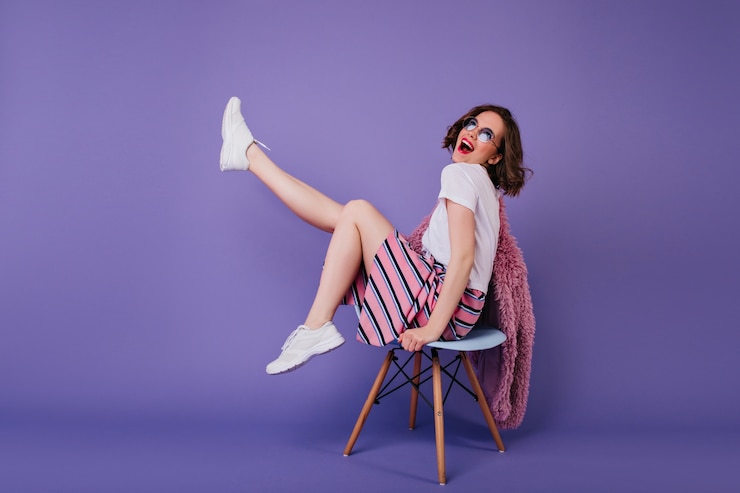 Foto grátis garota romântica em sapatos brancos, sentada na cadeira e rindo. tiro interno de mulher encaracolada despreocupada, posando na parede roxa com um sorriso sincero.