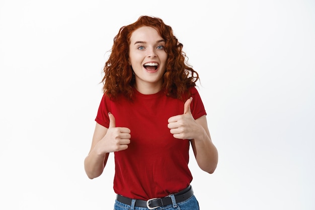 Foto grátis garota ruiva animada elogiando o bom trabalho, mostrando os polegares para cima e sorrindo satisfeita, acena em aprovação e diz que sim, dá uma resposta positiva, encostada na parede branca