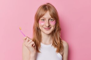 Foto grátis garota ruiva aplica manchas de hidrogel sob os olhos segura escova de dentes passa por procedimentos de beleza e higiene em sorrisos matinais agradavelmente isolados sobre fundo rosa cuida da pele e dos dentes