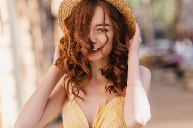 Garota ruiva bem-humorada em roupas amarelas, posando na rua borrada. Mulher ruiva refinada de chapéu, passar um tempo na cidade.