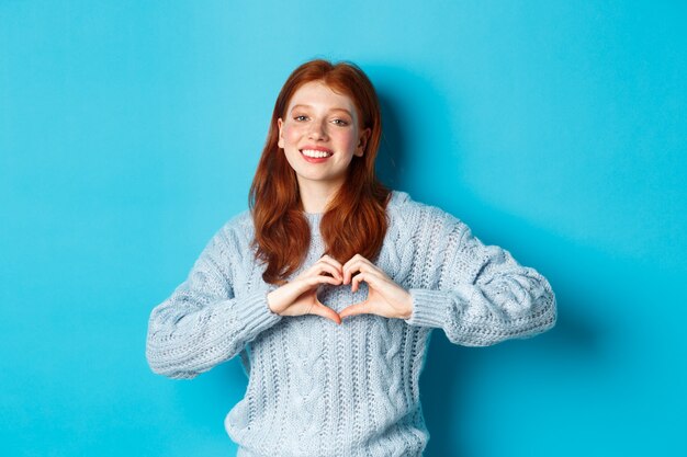 Garota ruiva bonita camisola mostrando o sinal do coração, eu te amo gesto, sorrindo para a câmera, em pé contra um fundo azul.