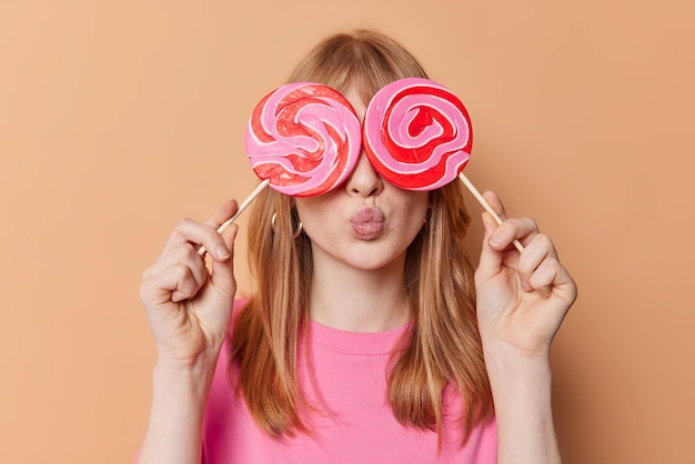 Foto grátis garota ruiva cobre os olhos com doces redondos de caramelo em palitos mantém os lábios dobrados tem vício em açúcar guloso usa camiseta casual isolada sobre fundo bege adolescente feminino com pirulitos