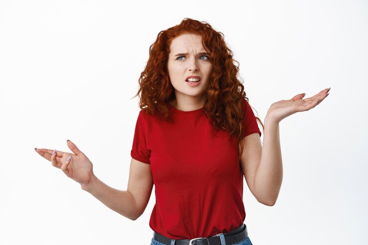 Foto grátis garota ruiva confusa não consegue entender algo, encolhendo os ombros e franzindo a testa confusa, olhando para o canto superior esquerdo perplexa, parada no branco