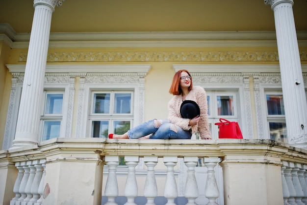 Garota ruiva de óculos e chapéu com bolsa vermelha posou perto de casa vintage