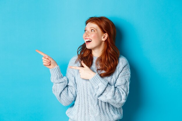 Garota ruiva empolgada de suéter, olhando e apontando o dedo para a esquerda, mostrando a oferta promocional ou logotipo, de pé sobre um fundo azul