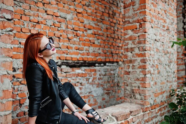 Garota ruiva estilosa em óculos de sol usa preto contra lugar abandonado com paredes de tijolos