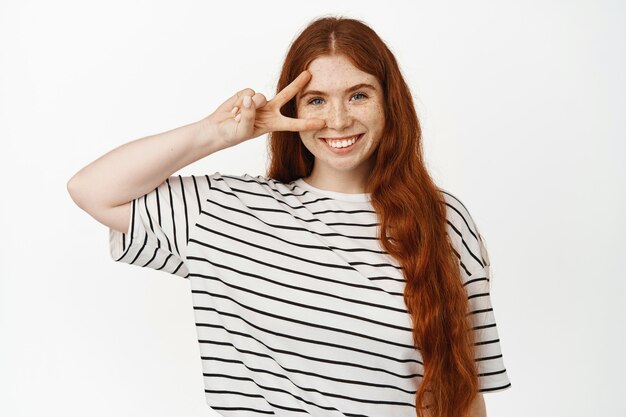 Garota ruiva feliz e positiva mostrando o símbolo da paz perto de limpar a pele sem maquiagem, sinal de V no olho, alegre de pé em t-shirt no estúdio branco.