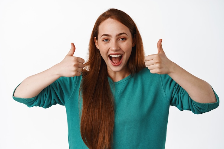 Foto grátis garota ruiva feliz e sorridente, mostrando os polegares em aprovação, satisfeita com algo legal, elogio e elogio, dê um feedback positivo sobre o branco.