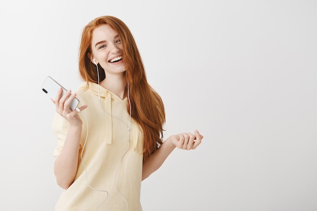 Garota ruiva feliz e sorridente ouvindo música em fones de ouvido e segurando um telefone celular