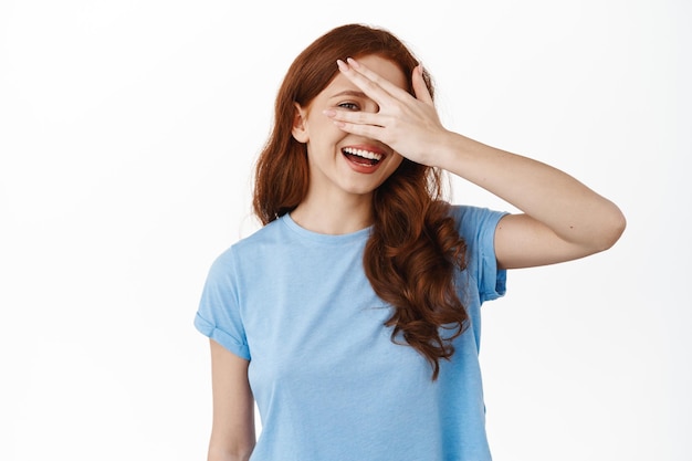 Foto grátis garota ruiva feliz escondendo o rosto, espiando por entre os dedos com cara alegre, sorrindo amplamente, de pé na camiseta contra fundo branco