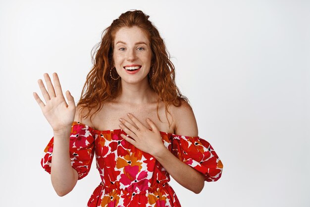 Garota ruiva feliz levantando a mão e colocando o braço no coração sorrindo amigável dizendo olá acenando para você de pé no vestido de verão contra fundo branco