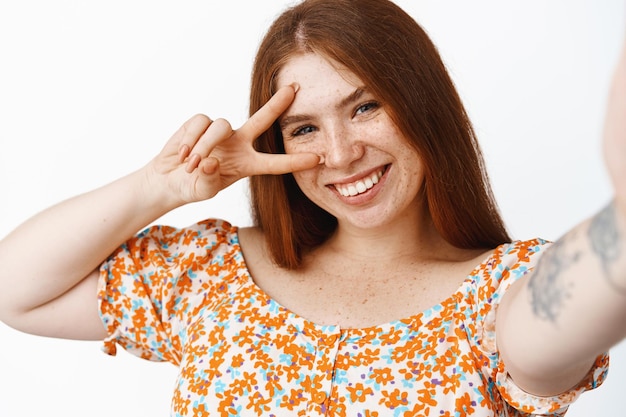 Garota ruiva feliz tomando selfie no celular posando com paz vsign e sorrindo em pé sobre fundo branco