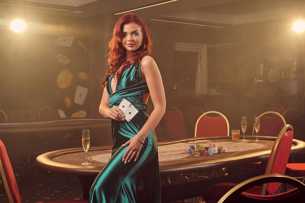 Garota ruiva sedutora em um longo vestido de cetim azul está sorrindo e posando de lado com dois ases na mão contra uma mesa de pôquer no cassino de luxo. Paixão, cartas, fichas, álcool, vitória, jogos de azar - é eu