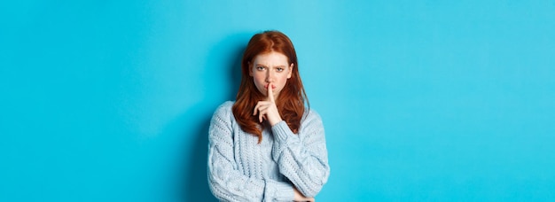 Foto grátis garota ruiva zangada silenciando para você mostrar que o gesto tabu proíbe falar em pé sobre o fundo azul i