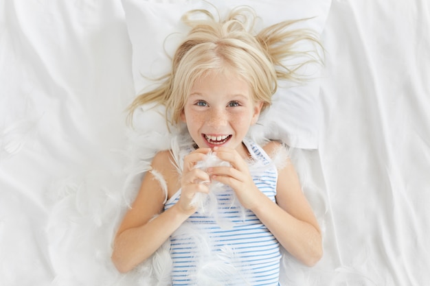 Garota sardenta bonita brincando com o irmão na cama, brigando com almofadas, pegando penas, tendo uma expressão feliz. loira sardenta menina brincando com penas deitado no travesseiro branco na cama