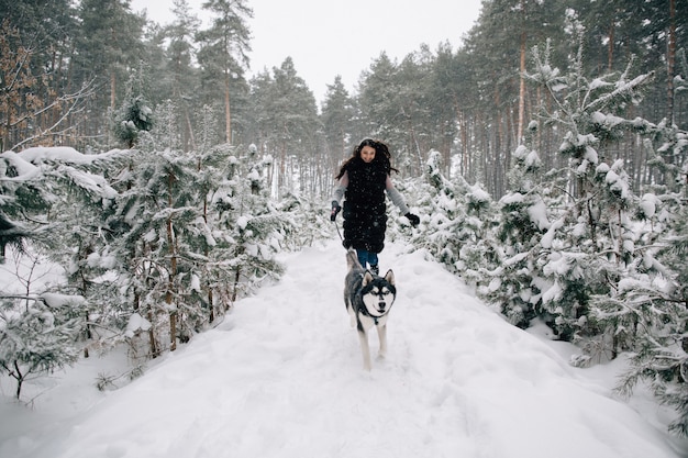 Garota se divertir com seu cachorro Husky na floresta de pinheiros de inverno nevado