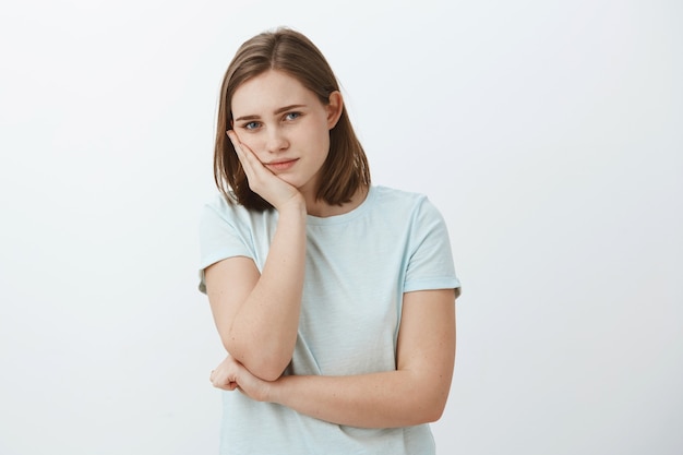 Garota se sentindo entediada com a rotina. Retrato de uma mulher europeia calma e bonita, inclinando a cabeça na palma da mão, olhando com um olhar cansativo e indiferente, sem ter nada para fazer em pé contra uma parede cinza