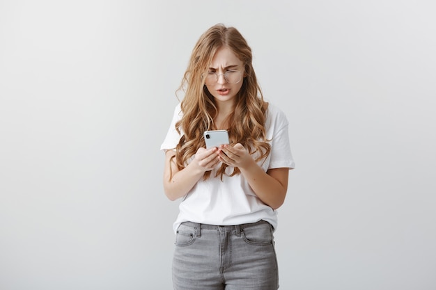 Foto grátis garota se sentindo estranha ao enviar mensagem para o número errado. retrato de uma jovem atraente com problemas de óculos, xingando enquanto franzia a testa e parecendo incomodado com a tela do smartphone, navegando na rede