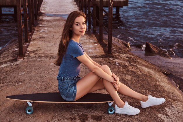 Garota sensual sentada em um skate no velho cais está desfrutando de um incrível clima nublado durante o pôr do sol.