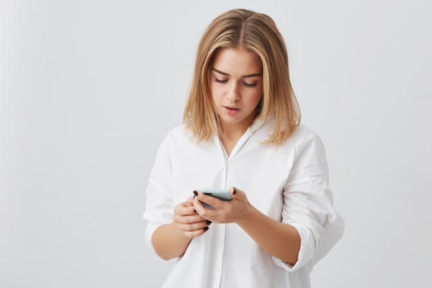 Garota séria com cabelo loiro, vestindo camisa branca, navegar na Internet no telefone celular, olhando para a tela com expressão concentrada, verificando e-mail contra parede cinza studio espaço cópia