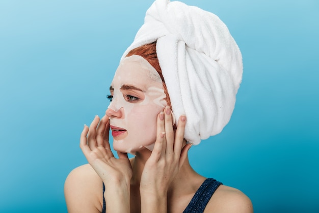 Garota sonhadora, aplicando a máscara facial isolada sobre fundo azul. Foto de estúdio de jovem com uma toalha na cabeça, olhando para longe.