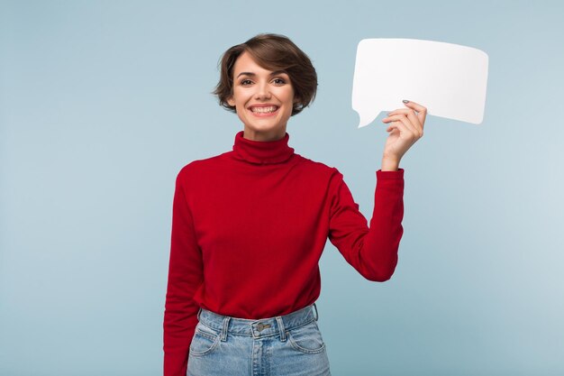 Garota sorridente atraente com cabelo curto escuro no suéter vermelho olhando alegremente na câmera segurando a forma de papel branco da mensagem na mão sobre fundo azul isolado