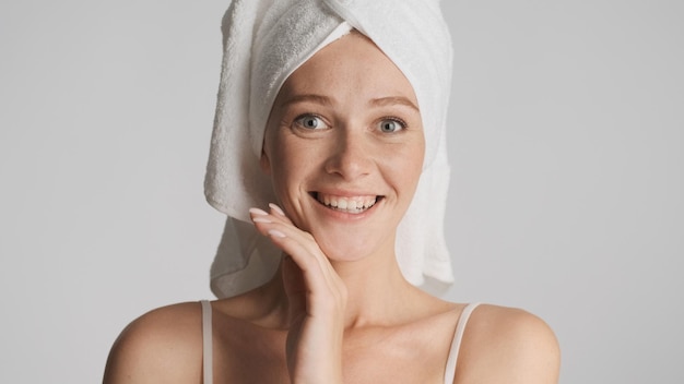 Garota sorridente atraente com toalha na cabeça olhando alegremente na câmera sobre fundo branco Conceito de beleza